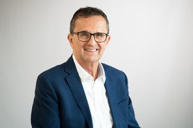 A smiling business man with short dark hair and black glasses wearing a white shirt and blue suit jacket